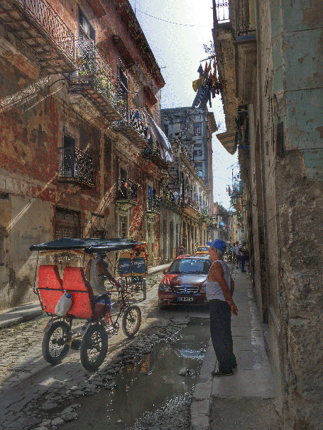photo of narrow Cuban city street as a man looks at a passing foot taxi
