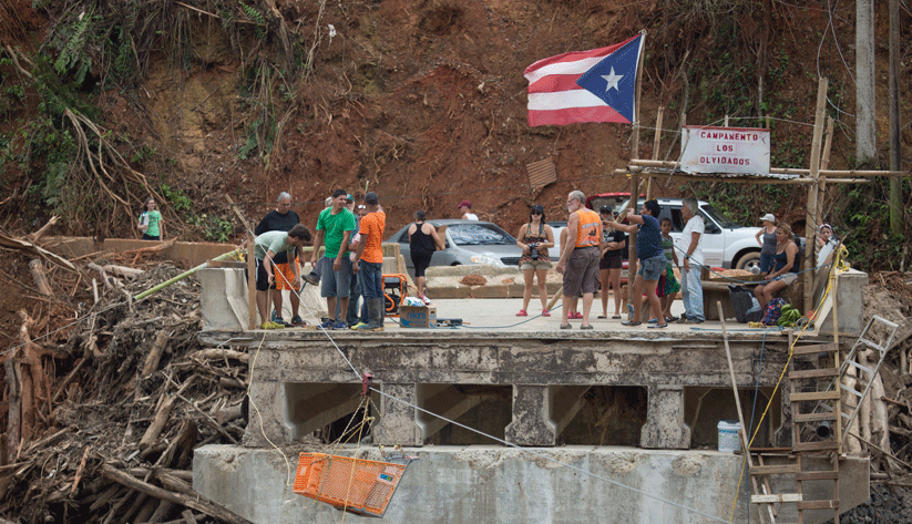 Villagers gather on bridge remnants to receive needed supplies. The village was cut off by the Rio Charco Abajo's flooding that made the village road and bridge impassable. A cable-pulley system ferries a shopping cart from shore-to-shore. Photo by Andrea Booher, courtesy of the Federal Emergency Management Agency