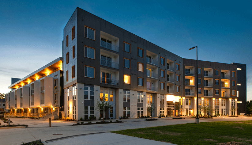 photograph of a newly-completed, five-story, mixed-use apartment building at dusk with its lights on